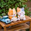 Two plush climbing chalk bags, Boba Dog (a corgi design) and Boba Cat (a calico design), sitting on a wooden bench alongside a vintage blue truck planter with succulents. Boba Dog holds a boba cup with a playful expression, and Boba Cat has a cheerful smile while holding another boba drink