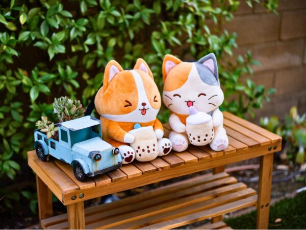 Two plush climbing chalk bags, Boba Dog (a corgi design) and Boba Cat (a calico design), sitting on a wooden bench alongside a vintage blue truck planter with succulents. Boba Dog holds a boba cup with a playful expression, and Boba Cat has a cheerful smile while holding another boba drink